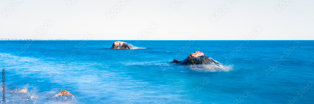 Zen-like seascape with two large glacial rocks and small stones in the corner. Blue ocean with white soft waves on the white sky background.