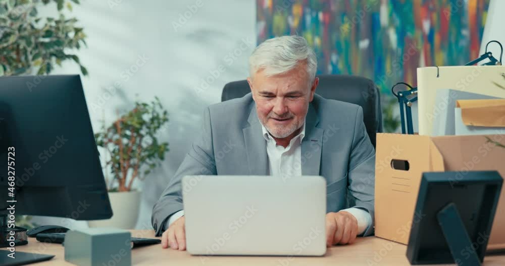 Man in suit with gray hair leads remote conversation from office, sitting at desk in front of laptop screen, waving hello to colleagues online meetings via webcam video chat talks about impressions Man in suit with gray hair leads remote conversation from office, sitting at desk in front of laptop screen, waving hello to colleagues online meetings via webcam video chat talks about impressions