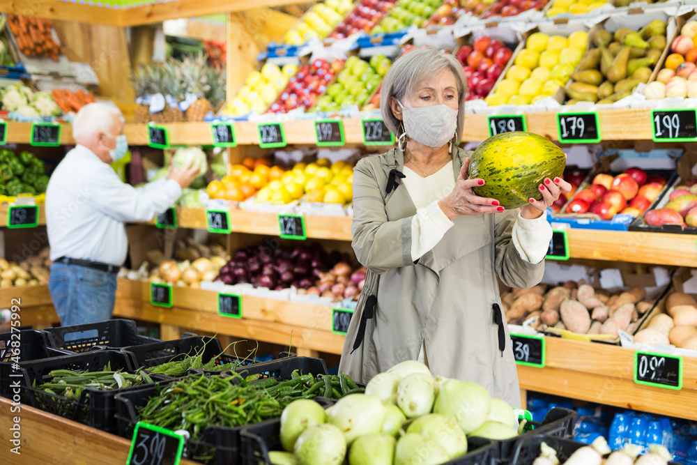 Woman in face mask choosing melon while standing in greengrocer. Old man shopping in background.