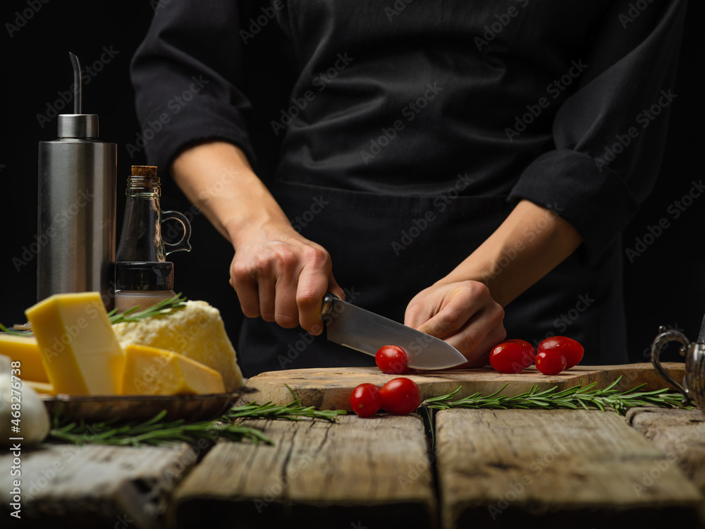 The chef is cutting tomatoes on a cutting board. Ingredients for making salad, sauce, pizza. Wooden texture, black background. Home cooking, hotel, restaurant, cafe, culinary blog.