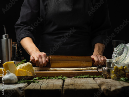 The chef rolls out the dough with a rolling pin on a cutting board. Ingredients for making pizza, pie, focaccia. Lots of facilities. Cookbook, cooking blog, instagram, advertising.