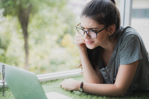 Beautiful young woman wearing eyeglasses lying near window on grass carpet mat engrossed in watching a movie or web series on laptop using internet connection at home