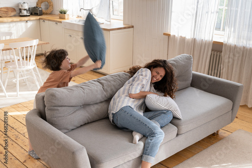Playful young mother has fun with preschool son fight pillows in living room. Active parent enjoy leisure time with small child. Funny family activities with mom and kid at home on lockdown or weekend photo