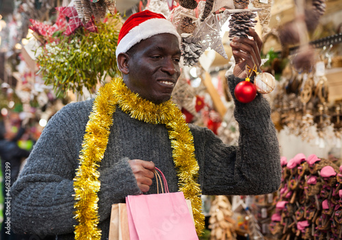 Cheerful African American in Santa hat looking for New Year decorations in traditional street market.