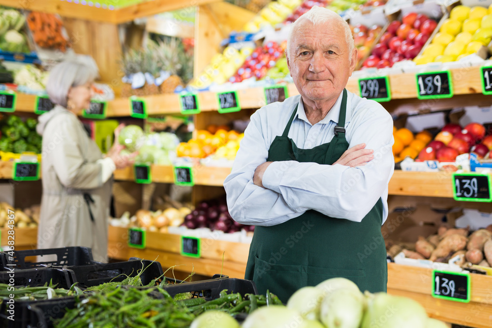 Old man supermarket worker standing in salesroom amongst shelves with vegetables and fruits and looking in camera. Woman making purchases in background.