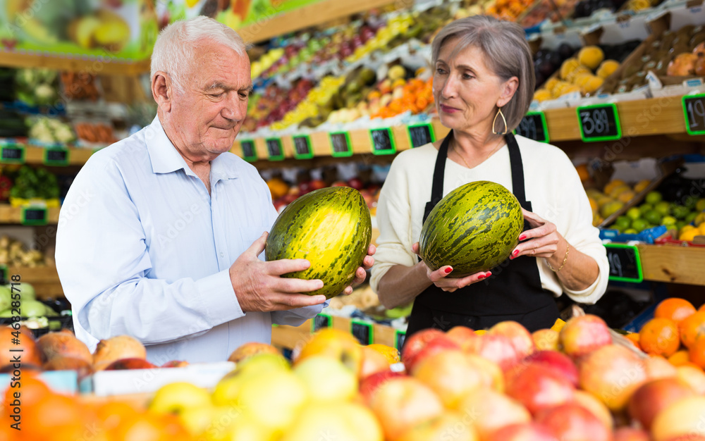 Mature woman greengrocer worker helping old man to choose fresh melon.