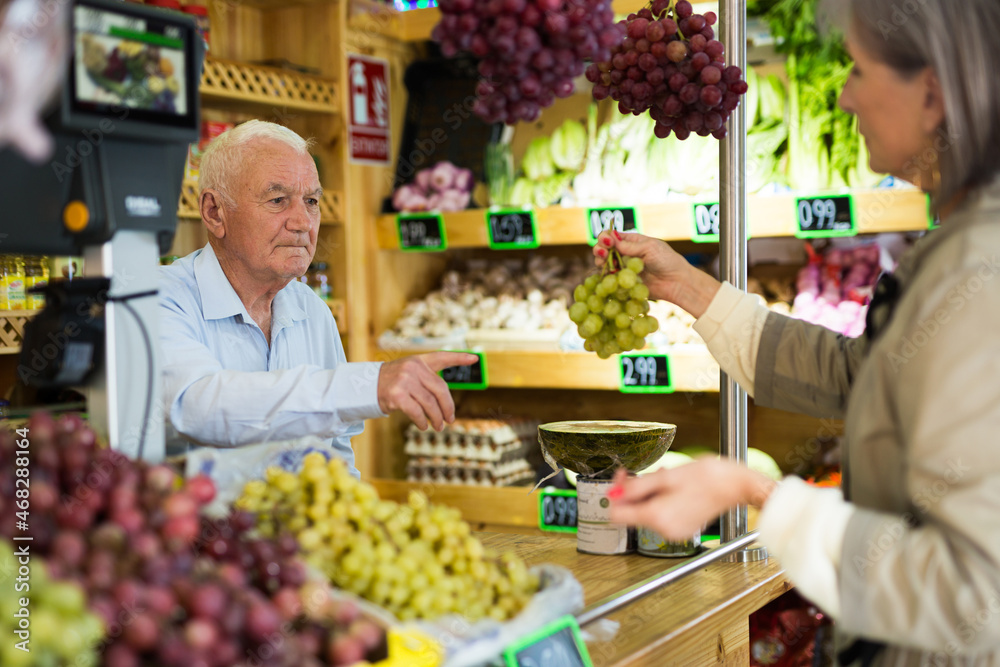 Old salesman standing at counter in greengrocer and serving mature woman who buying bunch of grape.