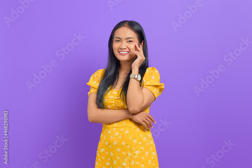 Smiling young Asian woman wearing yellow dress and looking confident on purple background