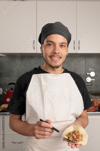 Young Latin American cook puts meat filling in empanada. As he cooks he looks at the camera smiling. Traditional South American food. Small entrepreneur works in his kitchen.