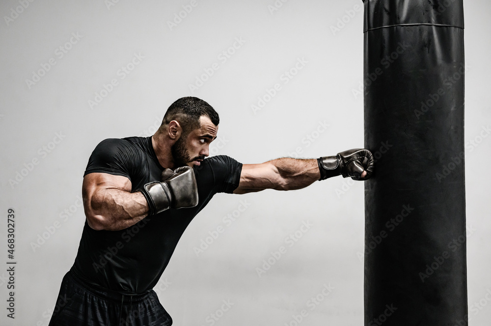 strong young caucasian bearded male in black sportswear kick punching bag with hands in boxing gloves on bright background in gym with copy space strong young caucasian bearded male in black sportswear kick punching bag with hands in boxing gloves on bright background in gym with copy space