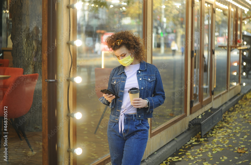 Beautiful curly haired woman in protective mask holding a phone in her hands and drinking coffee at outdoor cafes.