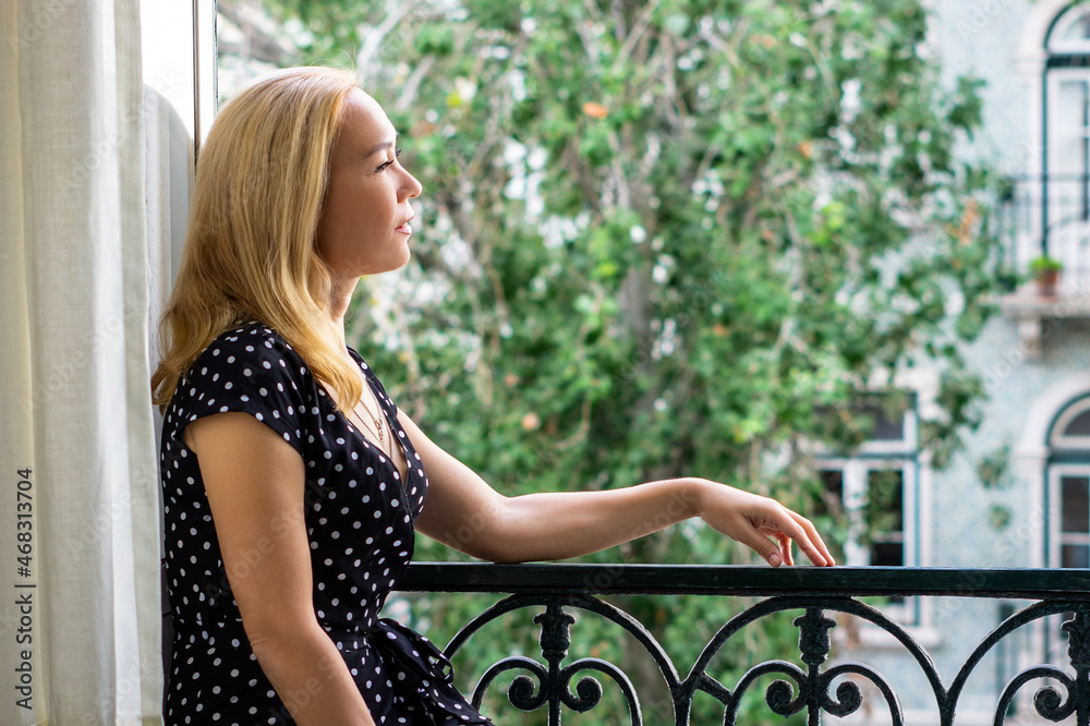 Happy blonde woman at opened window enjoying her day. Young woman female with blonde long hair in black dress with white polka dots in retro vintage style looks out of large window at summer sun day.
