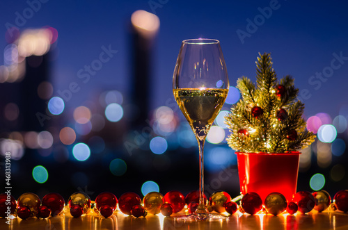 A glass of white wine that have Christmas tree decorated with bauble ornaments and lights on colorful city bokeh light background.