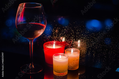 A glass of red wine with burning candles put beside window that have rain drop on dark background.