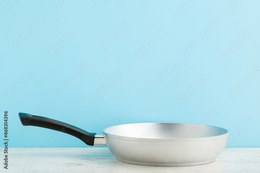 New aluminium frying pan on wooden table at light blue wall background. Pastel color. Closeup. Cooking concept. Empty place for text. Front view. New aluminium frying pan on wooden table at light blue wall background. Pastel color. Closeup. Cooking concept. Empty place for text. Front view.