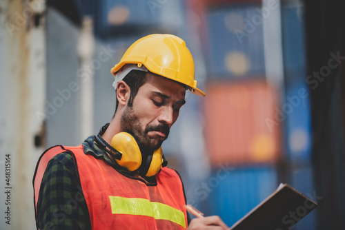 Handsome young male worker making note of shipping container with shipment and order number while wearing safety vest jacket and earphone at neck with hardhat helmet