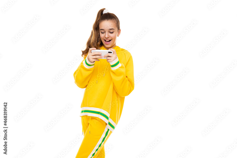Photo of a beautiful smiling cheerful cute girl with dark hair with a ponytail in a stylish yellow tracksuit with green stripes plays a mobile phone with a white case isolated on a white background Photo of a beautiful smiling cheerful cute girl with dark hair with a ponytail in a stylish yellow tracksuit with green stripes plays a mobile phone with a white case isolated on a white background