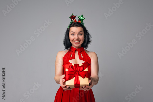 Happy young brunette woman in red dress posing on gray background and showing gift box decorated with red ribbon. Holidays, celebration and lifestyle concept