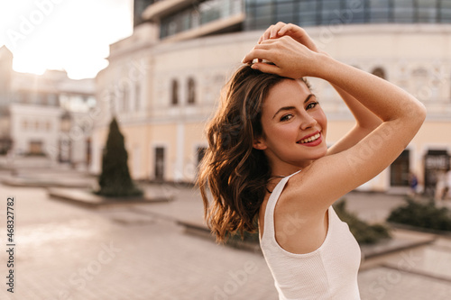 Joyful fair-skinned lady stands half sideways against background of city. Brown-haired woman with short hair holds her head with hands, smiling broadly at camera. © Look!