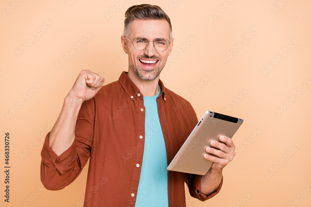 Photo of delighted middle-aged guy use gadget application raise fists up shopping sale isolated over pastel beige color background Photo of delighted middle-aged guy use gadget application raise fists up shopping sale isolated over pastel beige color background