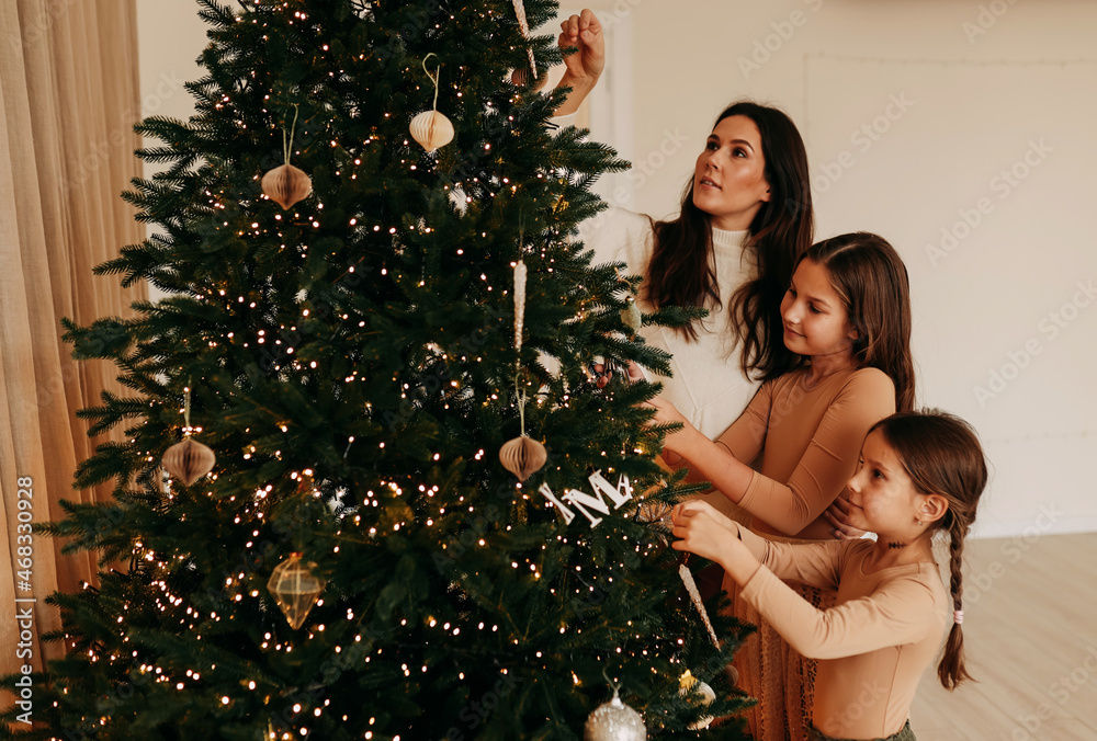 A happy mother with two children and daughters in casual clothes is decorating the Christmas tree together, preparing for the holiday, celebrating the new year in a cozy house. Selective focus