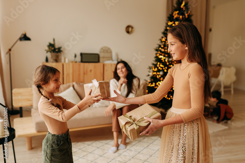 The concept of Christmas. Mom is sitting on the couch watching her little sister's daughters give each other gift boxes and wish each other a happy New year in the decorated cozy interior of the house
