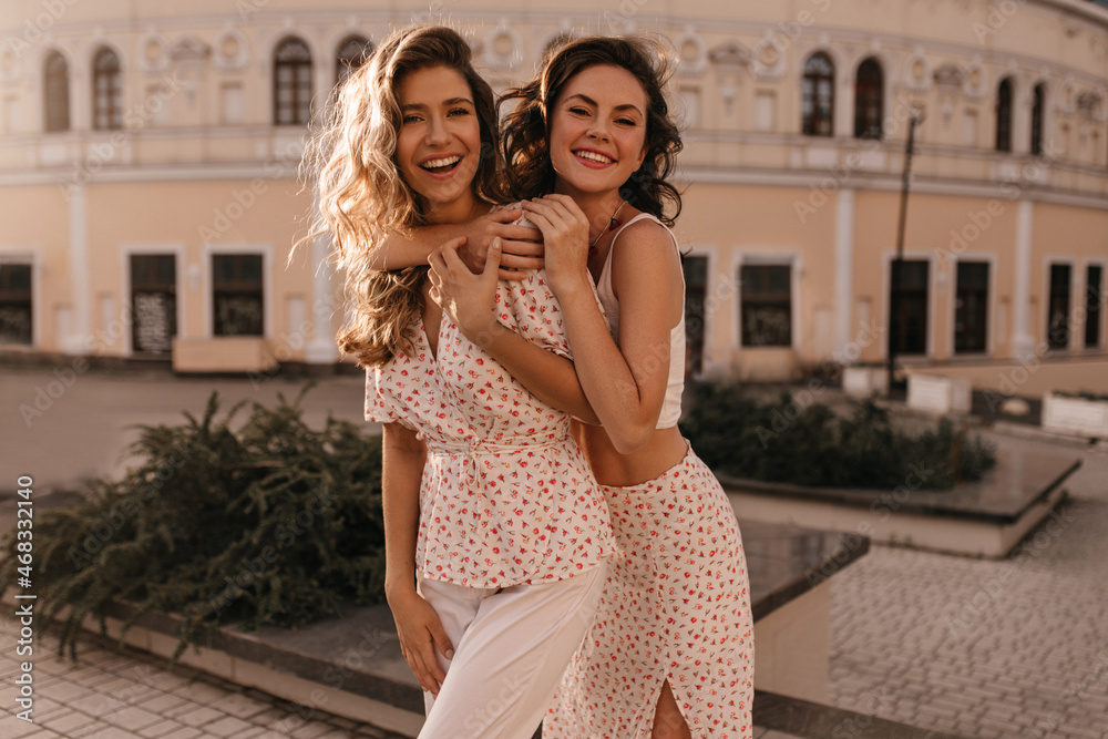 Fair-skinned pretty young lady hugs her girlfriend from back against backdrop of beige building. Teenagers with wavy loose hair are dressed in white summer clothes. Fair-skinned pretty young lady hugs her girlfriend from back against backdrop of beige building. Teenagers with wavy loose hair are dressed in white summer clothes.