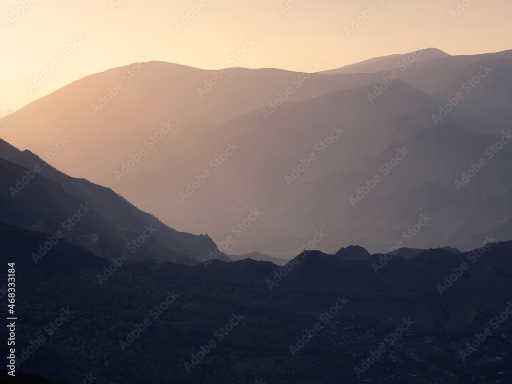 Scenic dawn mountain landscape with light fog in valley among mountains silhouettes under cloudy sky. Vivid sunset or sunrise scenery with low clouds in mountain valley in soft color.
