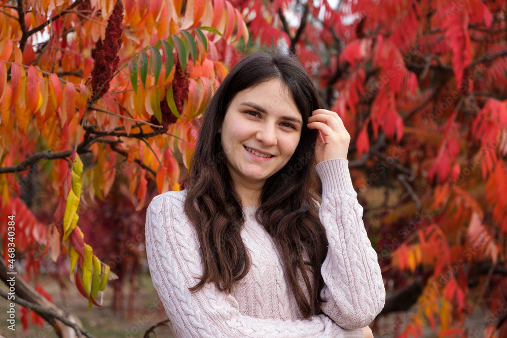 A 25-year-old brunette woman corrects her long hair and smiles against the background of red leaves in the fall