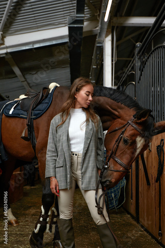 Smiling young woman standing inside a stable holding a saddle while preparing her chestnut horse for a ride