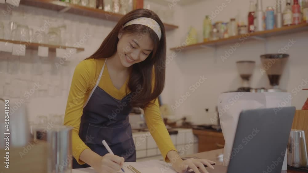 A beautiful Asian female employee of a coffee shop in an apron is working at her coffee shop or restaurant, checking bills, sales orders, using her laptop to work. A beautiful Asian female employee of a coffee shop in an apron is working at her coffee shop or restaurant, checking bills, sales orders, using her laptop to work.