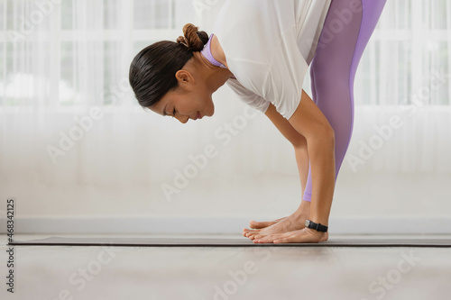 In the morning  an Asian woman is doing yoga  standing forward bend exercise  head to knees  uttanasana stance  working out  and wearing sportswear.