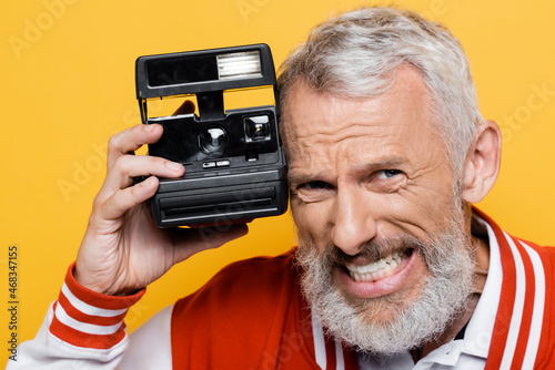middle aged man in bomber jacket holding black vintage camera and grinning isolated on yellow