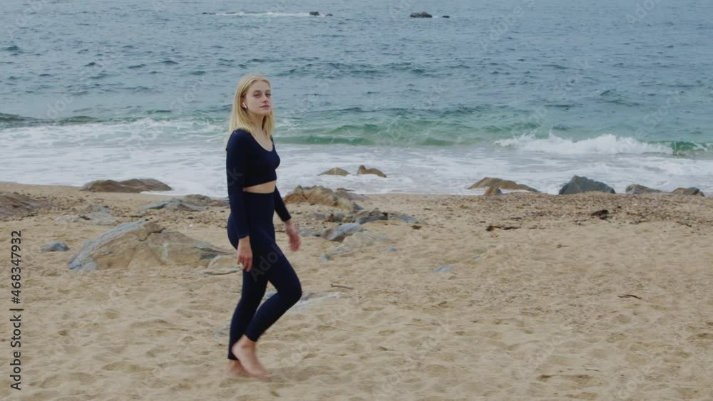 Young blond woman, fit and healthy, in sport outfit is walking on beach and looking on camera, enjoying active lifestyle and beautiful outdoor spot, Slow motion. Young blond woman, fit and healthy, in sport outfit is walking on beach and looking on camera, enjoying active lifestyle and beautiful outdoor spot, Slow motion.