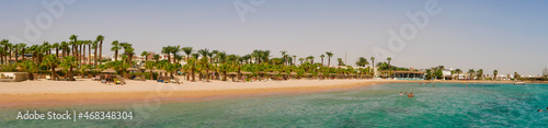 Hurghada, Egypt - September 22, 2021: Panoramic view of the sandy Egyptian beach with green palms. People sunbathe on sun loungers, swim in the Red Sea and snorkel against the blue sky. © lexosn