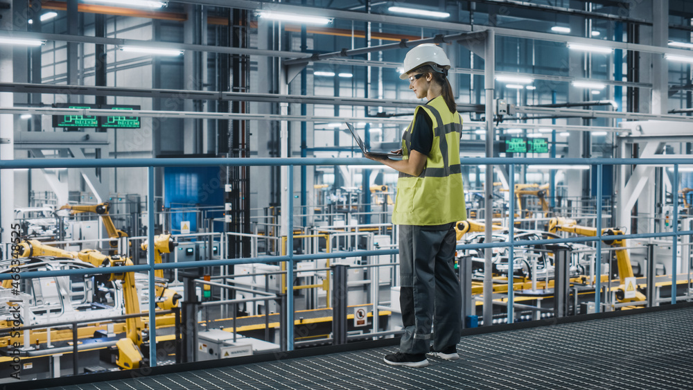 Female Car Factory Engineer in High Visibility Vest Using Laptop Computer. Automotive Industrial Manufacturing Facility Working on Vehicle Production with Robotic Technology. Automated Assembly Plant.