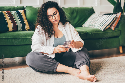 Curly haired plus size young woman wearing comfortable clothes in glasses clicks on smartphone screen sitting on floor mat in stylish room photo