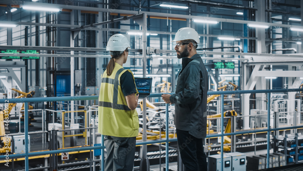 Male Specialist and Female Car Factory Engineer in High Visibility Vests Using Laptop Computer. Automotive Industrial Manufacturing Facility Working on Vehicle Production. Diversity on Assembly Plant.