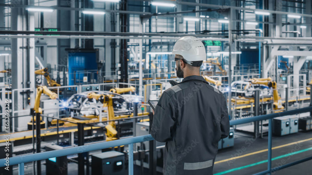 Car Factory Engineer in Work Uniform Using Tablet Computer. Automotive Industrial Manufacturing Facility Working on Vehicle Production with Robotic Arms Technology. Automated Assembly Plant. Car Factory Engineer in Work Uniform Using Tablet Computer. Automotive Industrial Manufacturing Facility Working on Vehicle Production with Robotic Arms Technology. Automated Assembly Plant.