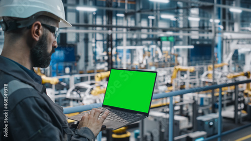 Car Factory Engineer in Work Uniform Using Laptop Computer with Green Screen Mockup Display. Working with Software at Automotive Industrial Manufacturing Facility Dedicated for Vehicle Production.