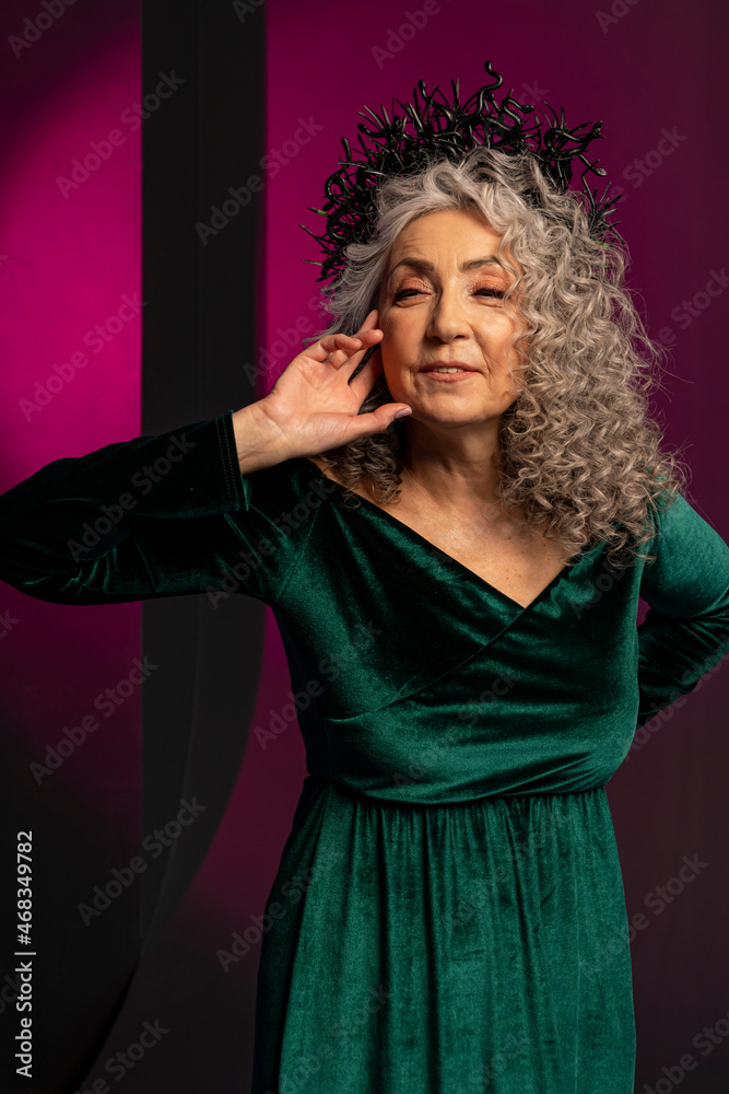 An elderly gray-haired elegant woman of 60-65 years old poses on a colored background, long curly hair. A stylish mature woman in a green dress with a deep neckline looks into the camera and smiles.