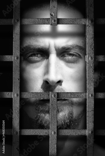 Caucasian adult bearded man looking through the bars of a prison cell photo