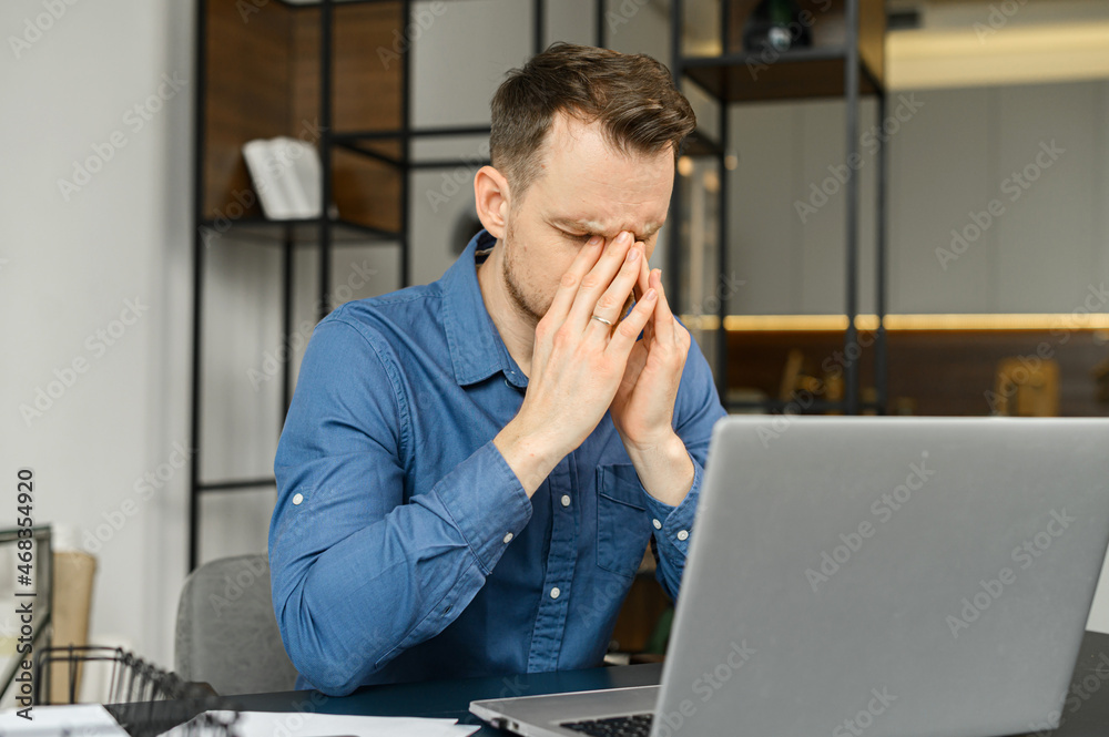 Fatigued guy using laptop for working, a man in smart casual wear is rubs the bridge of the nose, feels tired and burnout, sitting at the desk in modern office with eyes closed Fatigued guy using laptop for working, a man in smart casual wear is rubs the bridge of the nose, feels tired and burnout, sitting at the desk in modern office with eyes closed