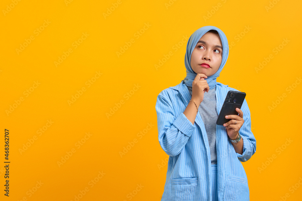 Pensive young Asian woman using mobile phone with fingers on chin over yellow background Pensive young Asian woman using mobile phone with fingers on chin over yellow background