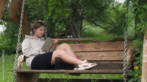 A young woman is sitting on a wooden swing and reading a book in nature. Outdoor recreation