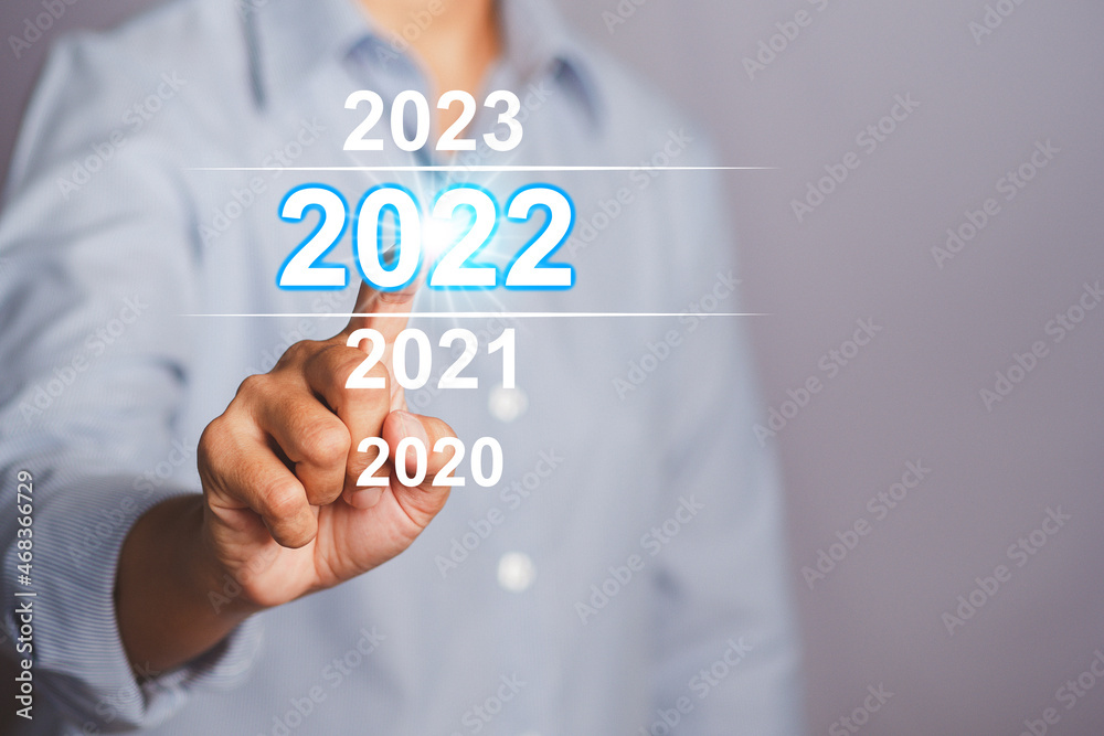 Welcome to the happy new year in 2022. Businessman in blue shirt pointing to digital 2022 calendar while standing with gray background in the studio. Business startup or goal to success Welcome to the happy new year in 2022. Businessman in blue shirt pointing to digital 2022 calendar while standing with gray background in the studio. Business startup or goal to success