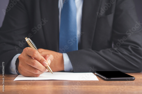 Close-up of a businessman holding a pen writing on paper with a blank screen mobile phone on a wooden desk in the office
