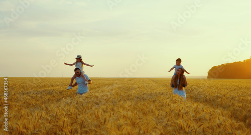 Parents holding little children on shoulders while walking in wheat field, children spreading arms playing airplanes, sky on background. Happy family together