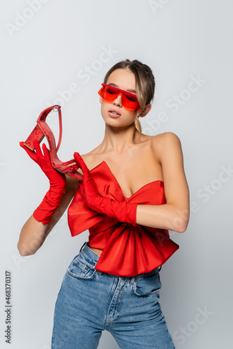 stylish model in red blouse, sunglasses and gloves holding red shoe isolated on grey photo