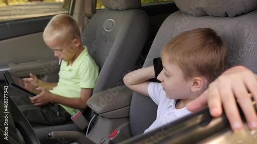 Two eight-year-old brothers are sitting behind the wheel of a car in the front with a phone and tablet.